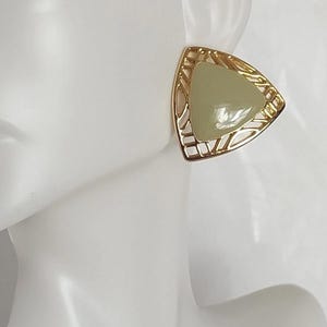 May include: A gold-toned, triangular earring with a pale green center stone. The gold frame features a geometric, openwork design. The earring is displayed on a white mannequin, showcasing its shape and color.
