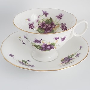 May include: A white teacup and saucer set with a floral violet design. The teacup has a scalloped rim and a gold trim. The saucer also has a scalloped edge and gold trim. The teacup is approximately 3 inches tall.