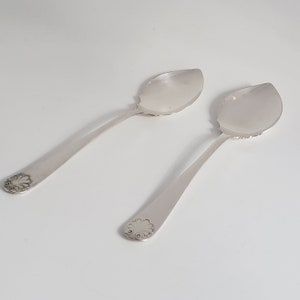 May include: Two silver serving spoons with a decorative floral design at the end of the handle. The spoons have a wide, oval bowl and a long, slender handle. The spoons are placed on a white surface.