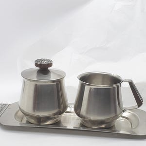 May include: A stainless steel creamer and sugar bowl set with a serving tray. The sugar bowl has a wooden knob on the lid. The creamer has a handle with a wooden accent. The tray has decorative edges.