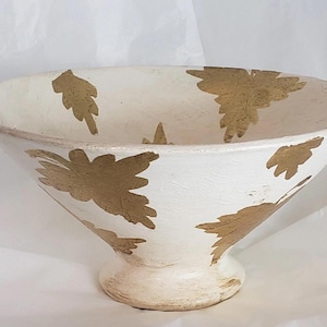 May include: White pedestal bowl with gold leaf pattern. The bowl has a wide, shallow shape and a rounded base. The gold leaf pattern is irregularly shaped and scattered across the surface of the bowl. The bowl is made of a textured material.