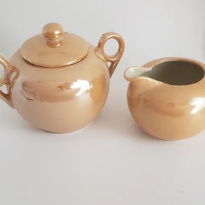 May include: A peach-colored ceramic tea set, including a teapot with a lid and a creamer. The teapot has a round shape with a handle and a spout. The creamer has a similar shape with a handle and a spout. The set has a glossy finish.