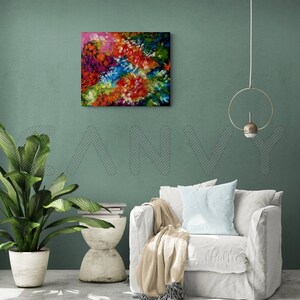 May include: Square abstract painting with a vibrant mix of colors including red, orange, blue, green, and pink. The artwork is displayed on a teal wall above a white armchair. A potted plant and side table are also visible.