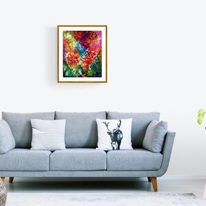 May include: A framed abstract art print with a gold frame. The artwork features a vibrant mix of colors, including red, yellow, blue, and green, creating a dynamic floral or landscape-inspired composition. The print is displayed above a gray sofa.