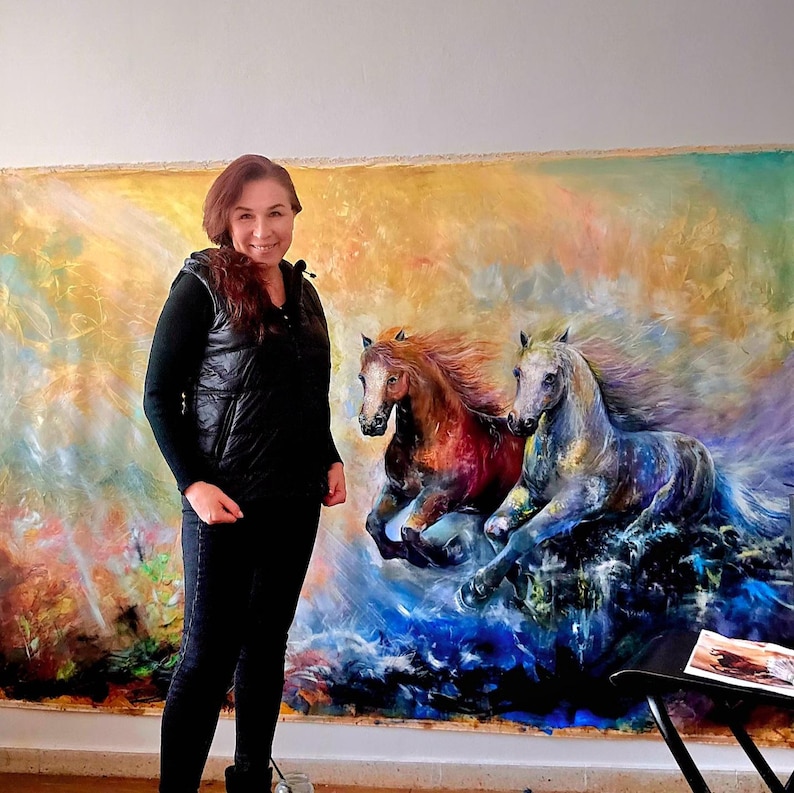 May include: A woman stands next to a large, colorful painting of two running horses. The horses are depicted with flowing manes and dynamic poses, set against a vibrant, abstract background with hues of orange, blue, and purple. The woman is wearing a black vest and black pants.