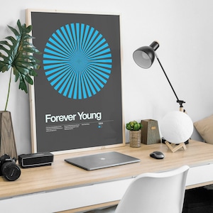 May include: A framed poster with a blue and white graphic design of a sunburst. The poster has the text "Forever Young" and the year "1984" printed on it. The poster is on a desk with a laptop, a lamp, and a moon lamp.