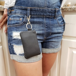 May include: A black leather key chain with a small zippered pouch attached. The pouch is hanging from a silver chain and is attached to a pair of denim shorts.