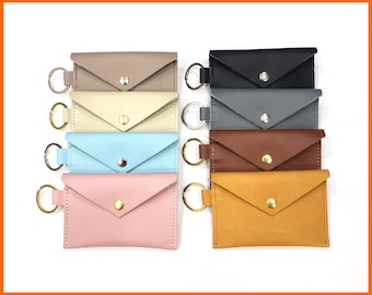 May include: Eight leather keychains with a flap closure and a metal ring. The keychains are in various colors, including beige, cream, light blue, pink, black, gray, brown, and tan.