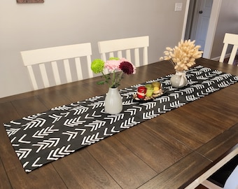 Peut inclure: Un chemin de table noir et blanc à chevrons avec des flèches blanches sur fond noir. Le chemin de table est sur une table en bois foncé avec deux chaises blanches et un vase de fleurs.