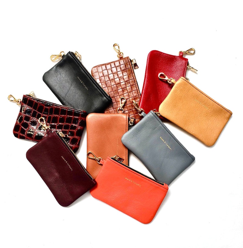 May include: Assortment of nine small leather pouches in various colors, including black, brown, red, orange, and tan. Each pouch has a gold zipper and a gold key ring attached. The pouches are arranged in a semi-circle on a white background.
