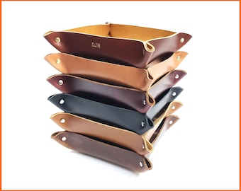 May include: A stack of leather valet trays in various colors, including brown, tan, and black. Each tray is rectangular with rounded corners and secured with metal rivets. The top tray has the initials "DJM" embossed.