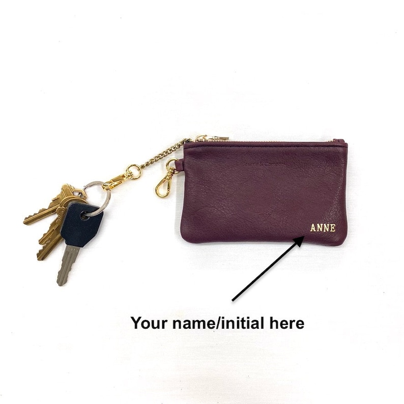 May include: A burgundy leather key chain with a gold chain and a zippered pouch. The pouch has a gold zipper and the text "ANNE" is embossed on the front.