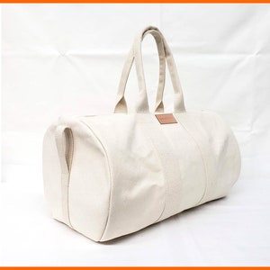 Może przedstawiać: Biała torba podróżna z płótna z brązowym skórzanym uchwytem i brązową skórzaną naszywką z przodu.