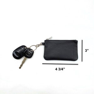 May include: Black leather key chain with a zipper closure. The keychain is 4 3/4 inches wide and 3 inches tall. It has a silver key ring with two car keys attached.