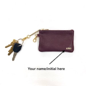 May include: A burgundy leather key chain with a gold chain and a zippered pouch. The pouch has a gold zipper and the text "ANNE" is embossed on the front.