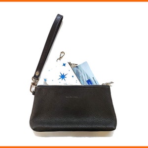 Könnte beinhalten: Eine schwarze Leder-Handgelenkstasche mit Reißverschluss und einem Handgelenkband. Die Handgelenkstasche ist offen und enthält eine weiße und blaue gemusterte Stofftasche und eine Kreditkarte.