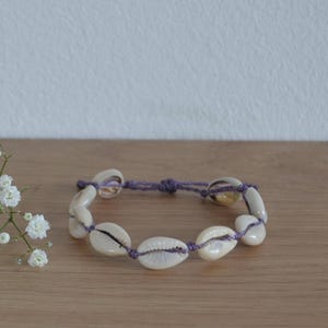 May include: A purple string bracelet with white seashells. The bracelet is tied with a knot and has a simple design.
