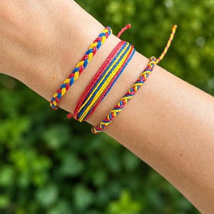 May include: A set of three bracelets featuring red, yellow, and blue cord. One bracelet is a braided design, the second consists of multiple parallel strands, and the third is a thinner braided cord. All three bracelets share the same colour palette.