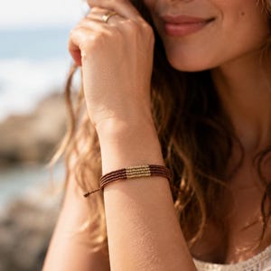 Op de afbeelding: Een bruine koordarmband met een centraal gedeelte van metallic goudkleurige kralen. De armband is gemaakt van meerdere strengen dun bruin koord die aan elkaar zijn geknoopt, met verstelbare uiteinden zichtbaar aan de zijkant van de pols.