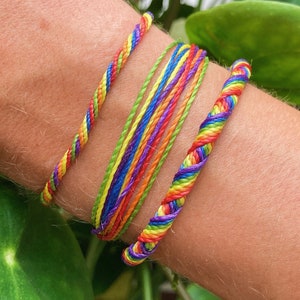 Peut inclure: Trois bracelets d'amitié aux couleurs de l'arc-en-ciel. L'un est torsadé, un autre tressé à plat, et le troisième est composé de fils parallèles. Les bracelets présentent des fils rouges, oranges, jaunes, verts, bleus et violets.