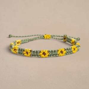 Può includere: Un braccialetto in macramè caratterizzato da un motivo ripetuto di girasoli gialli con centri marroni, intrecciato in un cordino verde salvia. Il braccialetto include una chiusura a nodo scorsoio regolabile con due piccole perline verdi alle estremità.