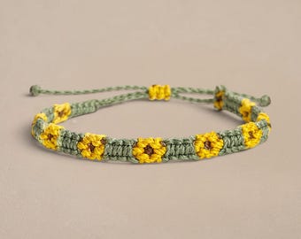 May include: A macrame bracelet featuring a repeating pattern of yellow sunflower motifs with brown centers, woven into a sage green cord. The bracelet includes an adjustable sliding knot closure with two small green beads at the ends of the cords.