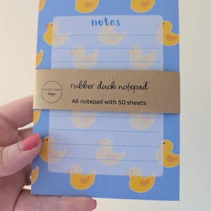 May include: A6 notepad with 50 sheets, featuring a blue background with yellow rubber duck illustrations. The notepad has lined pages with the word "notes" at the top. A brown paper band reads "rubber duck notepad".