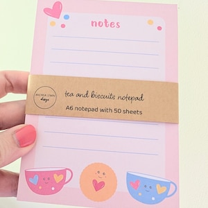 Pode incluir: Bloco de notas rosa A6 com o texto "notes" e "tea and biscuits notepad". O bloco de notas apresenta ilustrações de chávenas de chá e um biscoito. O bloco de notas tem 50 folhas.