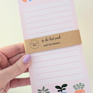 May include: A light pink to-do list pad with horizontal lines and a brown paper band labelled "to do list pad with 50 sheets" and "Andrea Lemin design". The pad features blue "to do" text, small heart and flower icons, and three potted plant illustrations.