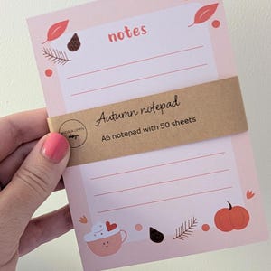 May include: A light pink A6 notepad with 50 sheets, featuring a brown paper belly band labelled Autumn notepad. The notepad has lined pages decorated with illustrations of leaves, a pumpkin, and a mug with a heart, and the word notes printed.