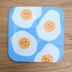 以下が含まれることがあります： 青い正方形のコースターに、黄色の卵黄と笑顔の顔をした白い目玉焼きの模様が4つ描かれています。