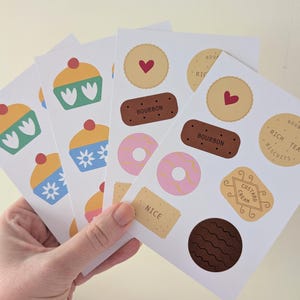 May include: A hand displays several cards featuring illustrations of baked treats. The designs include cupcakes, biscuits, and donuts. Some biscuits have the word "NICE" printed on them.