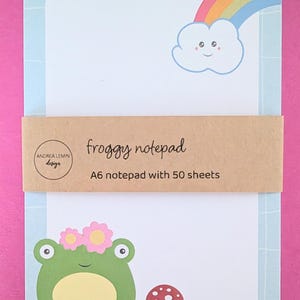 May include: A rectangular A6 notepad with 50 sheets, featuring a light blue grid border, a smiling cloud with a rainbow, a green frog with pink flowers, and a red mushroom. A brown paper band displays the text froggy notepad and Andrea Lemin design.