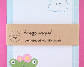 Könnte beinhalten: Ein rechteckiger A6-Notizblock mit 50 Blatt, hellblauem Gitterrand, einer lächelnden Wolke mit Regenbogen, einem grünen Frosch mit rosa Blumen und einem roten Pilz. Ein braunes Papierband zeigt den Text froggy notepad und Andrea Lemin design.