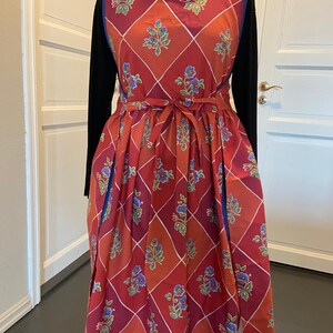 Könnte beinhalten: Eine rot-orangefarbene Schürze mit floralem Muster, Rautenmuster und blauem Besatz. Die Schürze hat eine Bindung an der Taille und ein blaues Blumenmuster. Die Schürze wird auf einer Schneiderpuppe präsentiert.