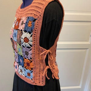 Könnte beinhalten: Eine ärmellose Häkelweste mit einem Vorderteil aus bunten Granny Squares in Blau-, Orange-, Rosa- und Brauntönen. Die Weste hat einen einfarbigen terrakottafarbenen Rand und seitliche Bindebänder, die das Vorder- und Rückenteil miteinander verbinden.