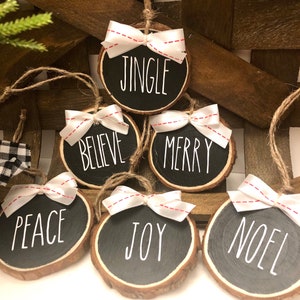 May include: Six round wood slice ornaments with white ribbon bows and red stitching. Each ornament has a different Christmas word written in white on a black background: Jingle, Believe, Merry, Peace, Joy, Noel.
