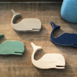 May include: Four wooden whale figurines in different colors: light gray, teal, white, and dark blue. The whales are all facing the same direction and are arranged in a semi-circle.