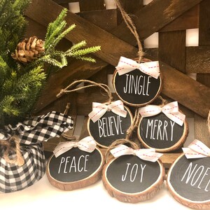 May include: Six round wood slice ornaments with white ribbon bows and red stitching. Each ornament has a different Christmas word written in white on a black background: Jingle, Believe, Merry, Peace, Joy, Noel.