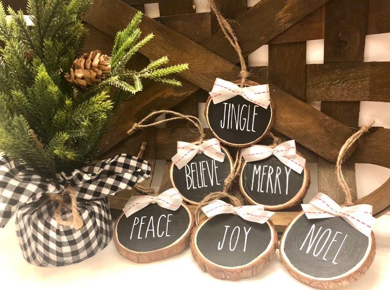 May include: Six round wood slice ornaments with white ribbon bows and red stitching. Each ornament has a different Christmas word written in white on a black background: Jingle, Believe, Merry, Peace, Joy, Noel.