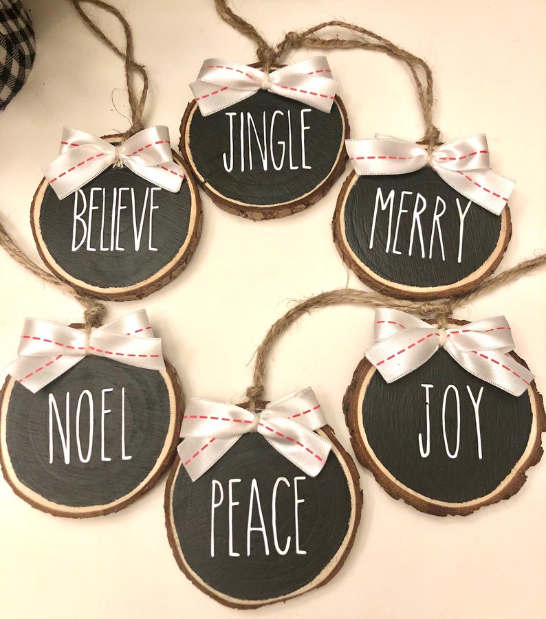 May include: Six round wood slice ornaments with white ribbon bows and black painted text. The ornaments read "Jingle", "Believe", "Merry", "Noel", "Joy", and "Peace".