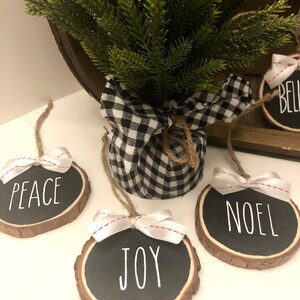 May include: Four round wood slice ornaments with black chalkboard paint and white lettering. The ornaments say "Peace", "Joy", "Noel", and "Believe". Each ornament has a white ribbon with red stitching tied around it.