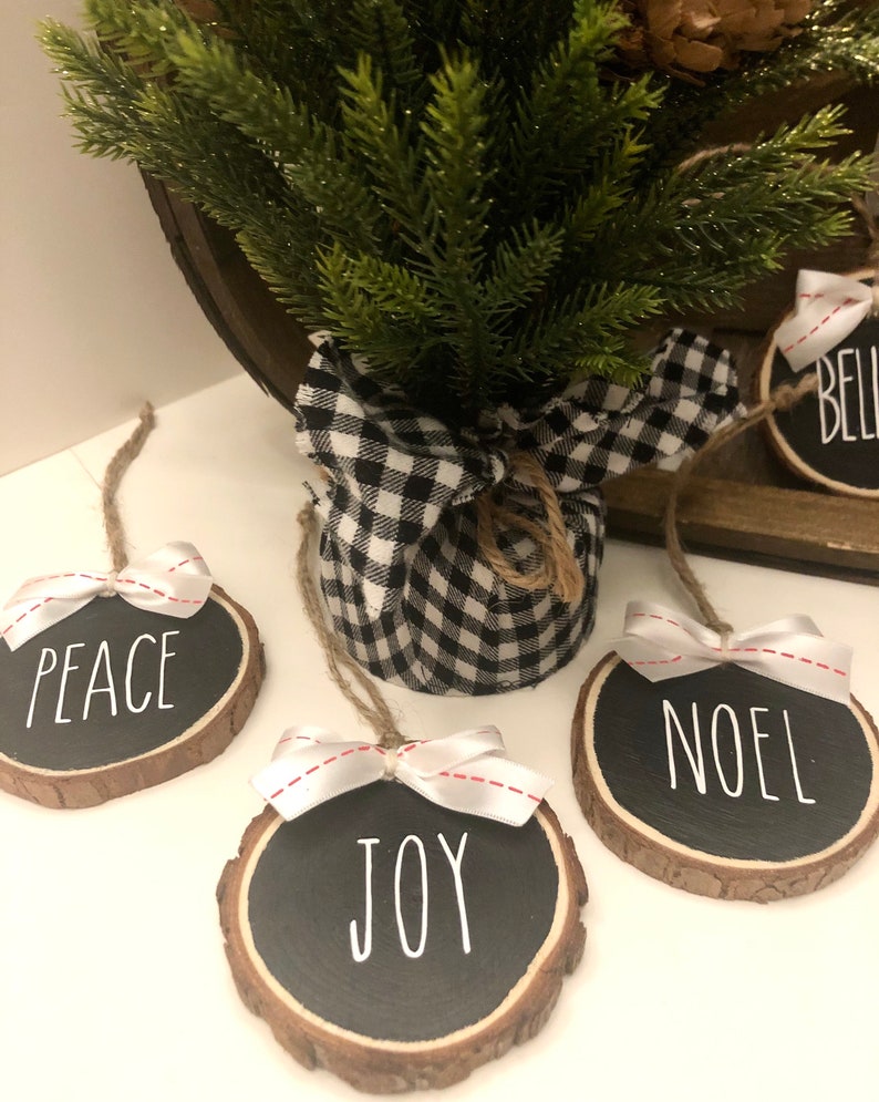 May include: Four round wood slice ornaments with black chalkboard paint and white lettering. The ornaments say "Peace", "Joy", "Noel", and "Believe". Each ornament has a white ribbon with red stitching tied around it.