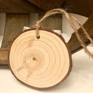 May include: A round wooden slice with a natural wood grain pattern and a hole drilled in the top. The slice is attached to a piece of twine, making it suitable for hanging as an ornament or tag.