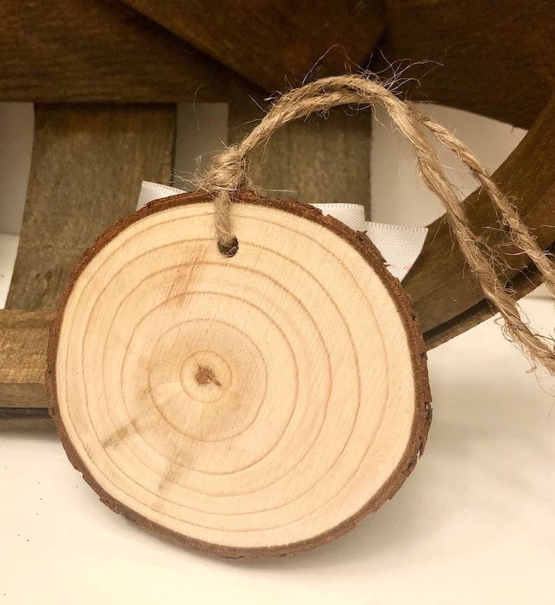 May include: A round wooden slice with a natural wood grain pattern and a hole drilled in the top. The slice is attached to a piece of twine, making it suitable for hanging as an ornament or tag.