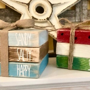May include: Two sets of stacked wooden blocks with painted designs. The first set is blue, brown, and white with the words "Sandy", "Salty", and "Happy" and an anchor. The second set is red, white, and green with the words "Hello", "Sweet", and "Summer".
