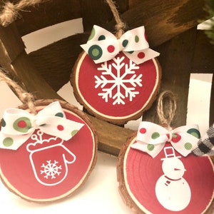 May include: Three red wooden ornaments with white designs. The ornaments are decorated with a snowflake, a mitten, and a snowman. Each ornament has a white ribbon with red and green polka dots.