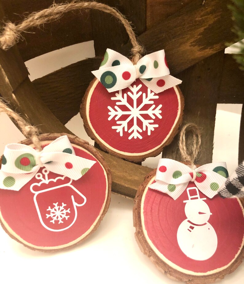 May include: Three red wooden ornaments with white designs. The ornaments are decorated with a snowflake, a mitten, and a snowman. Each ornament has a white ribbon with red and green polka dots.