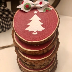 May include: A stack of five wooden round ornaments painted red with a white silhouette of a Christmas tree on the top ornament. The top ornament has a white ribbon with red and green polka dots tied around it.