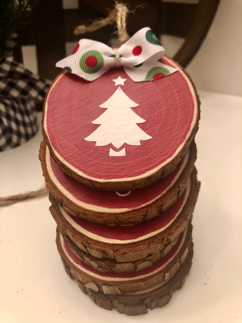 May include: A stack of five wooden round ornaments painted red with a white silhouette of a Christmas tree on the top ornament. The top ornament has a white ribbon with red and green polka dots tied around it.